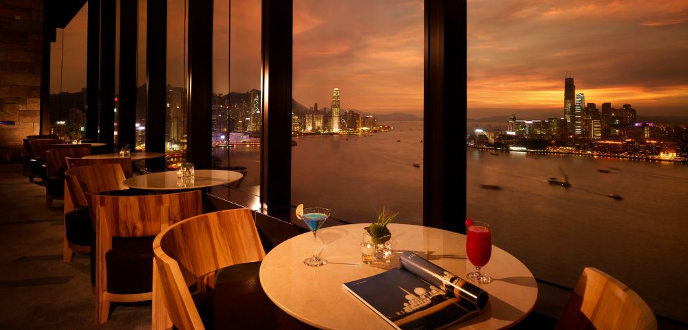 Upscale restaurant with large windows overlooks a harbor at sunset. Two tables have drinks, a magazine, and elegant décor, creating a warm, sophisticated ambiance.