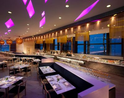 Modern restaurant interior with purple ceiling lights, sleek tables, and a buffet along large windows. Warm ambiance with a stylish, elegant feel.