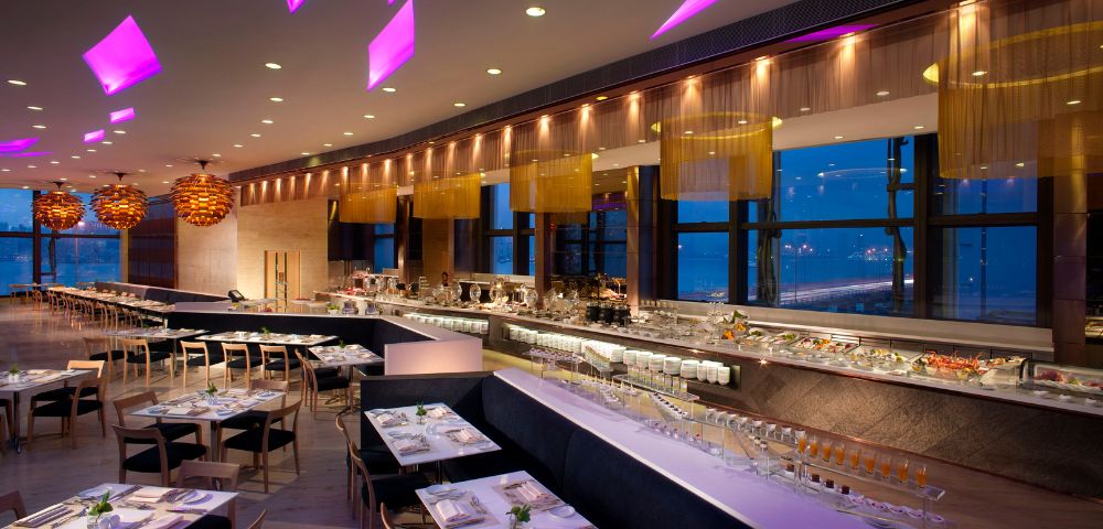 Modern restaurant interior with purple ceiling lights, sleek tables, and a buffet along large windows. Warm ambiance with a stylish, elegant feel.