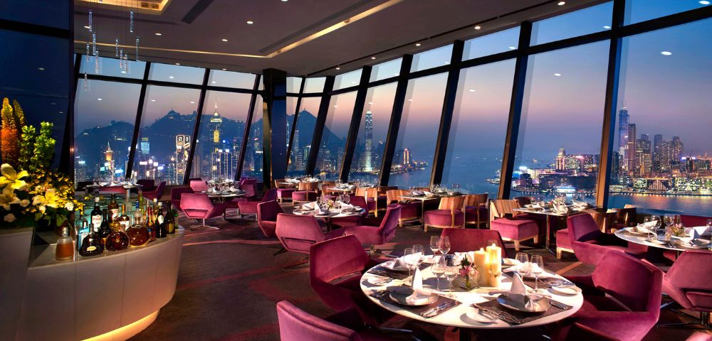 Luxurious restaurant interior with purple chairs and candlelit tables by large windows. Offers panoramic city skyline view at sunset, creating an elegant ambiance.