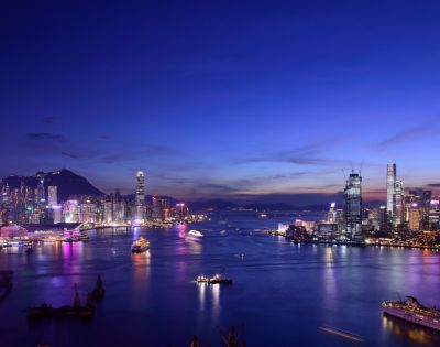 Vibrant cityscape at dusk with illuminated skyscrapers and colorful lights reflecting on a calm harbor, under a deep blue, twilight sky.
