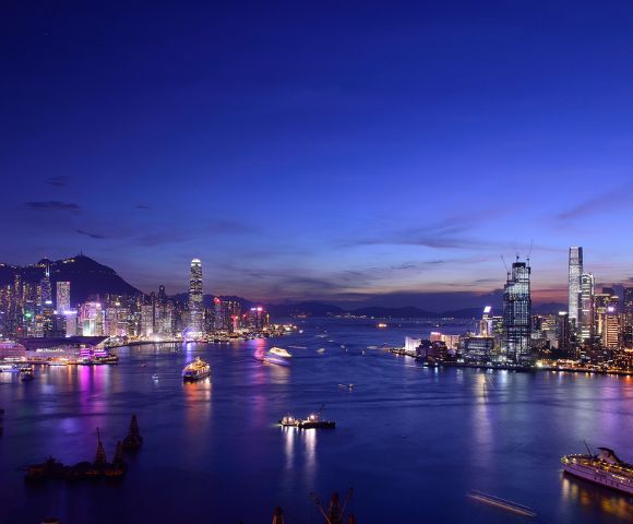 Vibrant cityscape at dusk with illuminated skyscrapers and colorful lights reflecting on a calm harbor, under a deep blue, twilight sky.