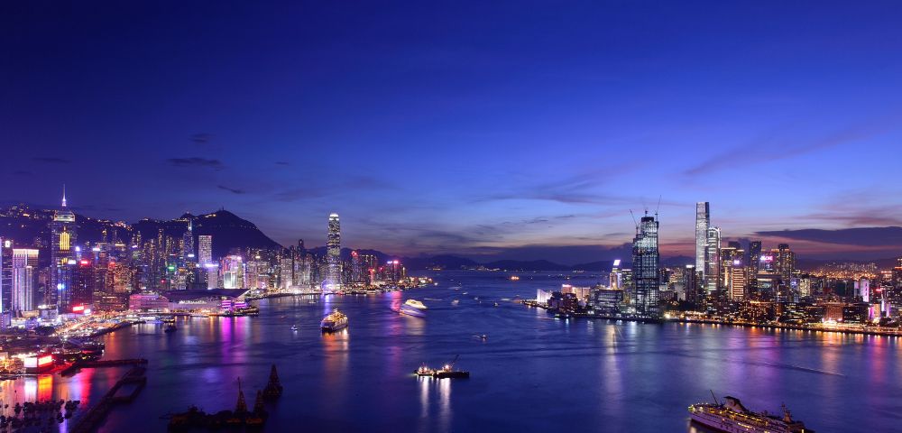 Vibrant cityscape at dusk with illuminated skyscrapers and colorful lights reflecting on a calm harbor, under a deep blue, twilight sky.