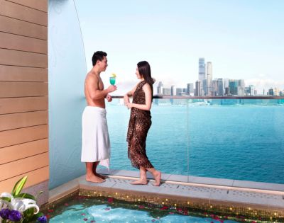 A man and woman stand on a balcony overlooking a city skyline and ocean. The man is in a towel, and the woman in a dress; both hold drinks, conveying a relaxed, luxurious vibe.