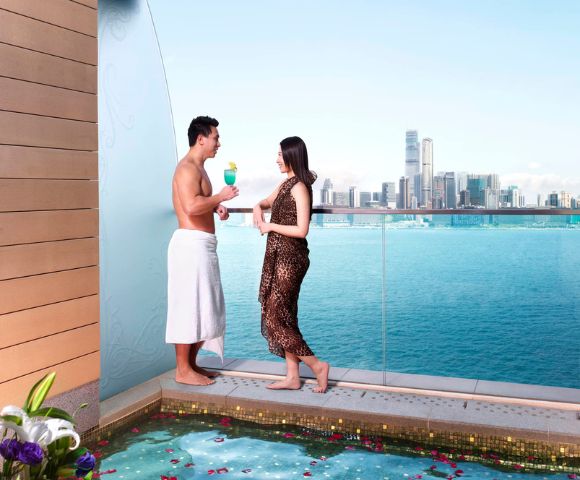 A man and woman stand on a balcony overlooking a city skyline and ocean. The man is in a towel, and the woman in a dress; both hold drinks, conveying a relaxed, luxurious vibe.