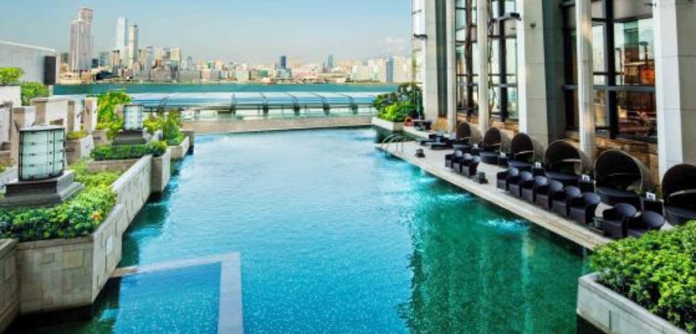 Luxurious rooftop pool overlooking a city skyline. Modern architecture, lounge chairs, greenery, and a serene, sunny atmosphere create a sense of relaxation.