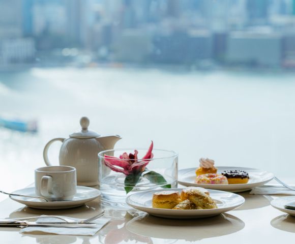 Elegant tea set on a table with pastries, teapot, and flower vase. Background shows a serene cityscape and water view, evoking a calm ambiance.