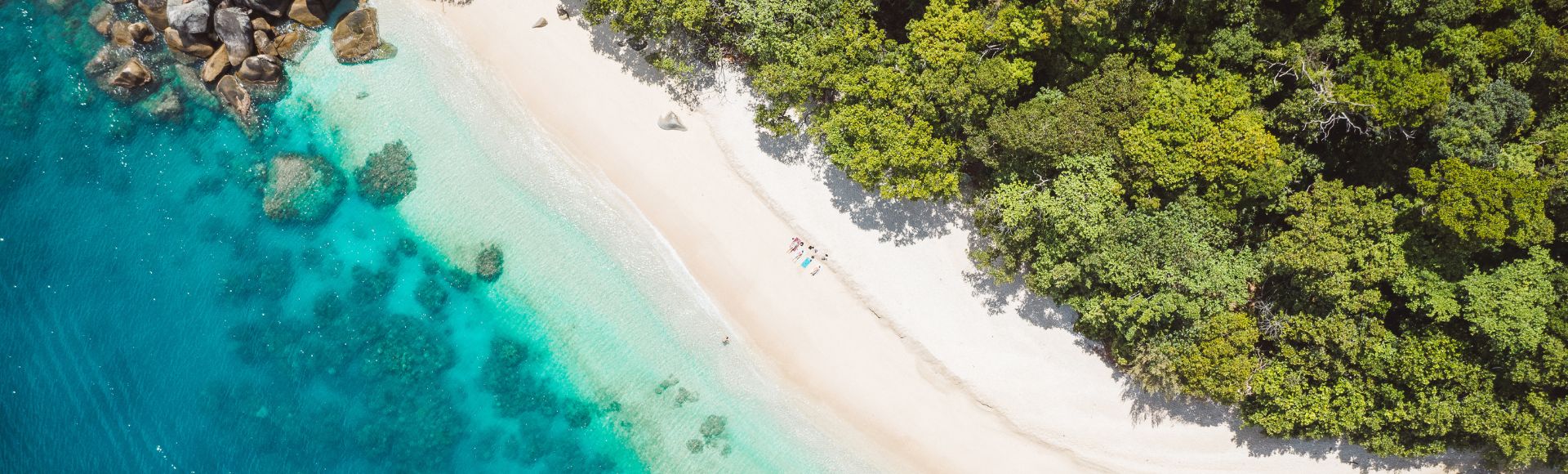 Aerial view of a secluded beach in Cairns with white sand, turquoise waters, and lush greenery, featuring a few people enjoying the serene environment.