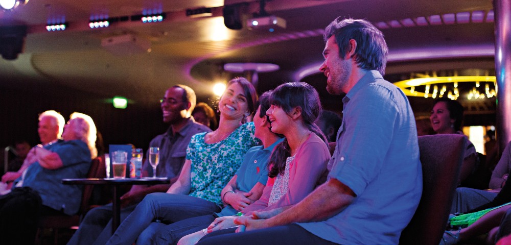 Audience members sit close together in a dimly lit theater, smiling and laughing. The mood is relaxed and joyful, with drinks on the table.