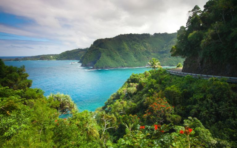 Lush green cliffs overlook a vibrant turquoise sea, with winding roads cutting through the scenic landscape under a partly cloudy sky.