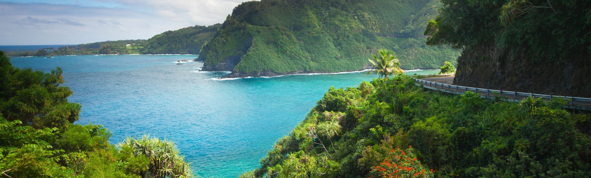 Lush green cliffs overlook a vibrant turquoise sea, with winding roads cutting through the scenic landscape under a partly cloudy sky.