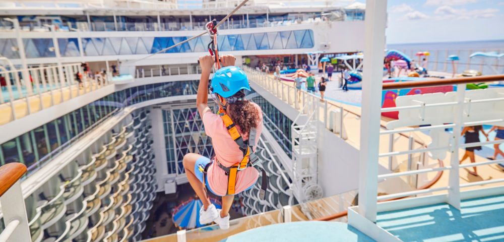 A person wearing a blue helmet and harness zip-lining over a cruise ship deck, with onlookers and vibrant activities in the background, conveying excitement and adventure.