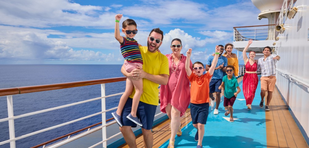 A joyful group of people on a cruise ship deck, dressed in colorful summer clothes, some with raised arms. The ocean and sky are in the background.