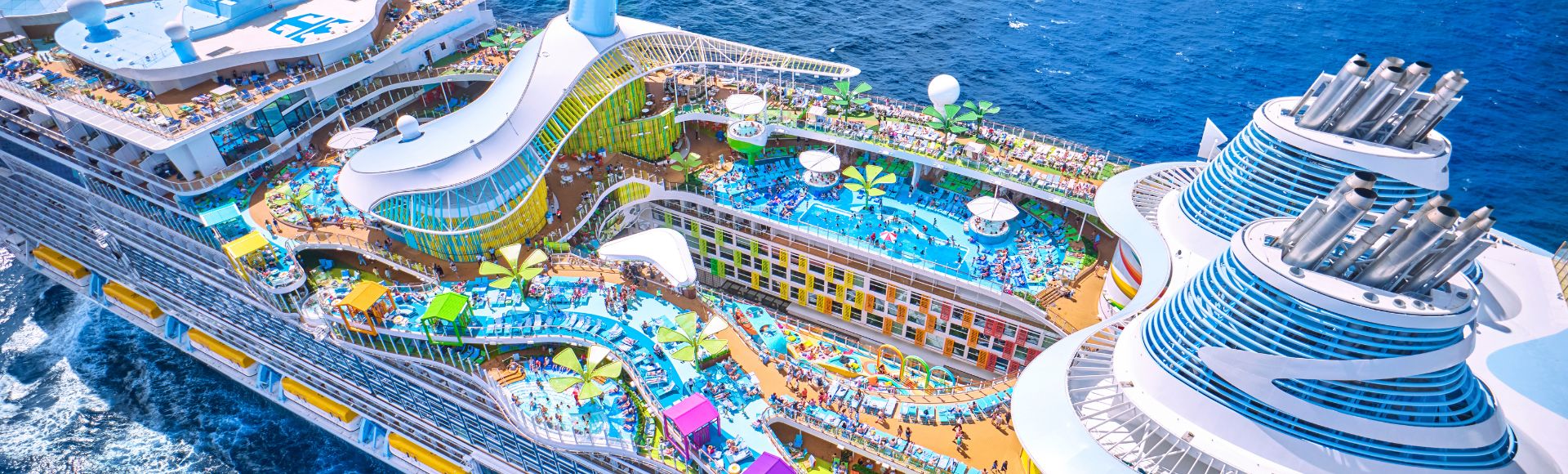 Aerial view of a colorful cruise ship deck with multiple pools, water slides, and lounging areas. The ship sails through vibrant blue ocean waters.