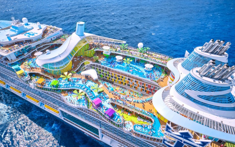 Aerial view of a colorful cruise ship deck with multiple pools, water slides, and lounging areas. The ship sails through vibrant blue ocean waters.