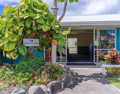 A small departure lounge entrance surrounded by lush green plants and colorful flowers on a sunny day, creating a welcoming, tropical atmosphere.