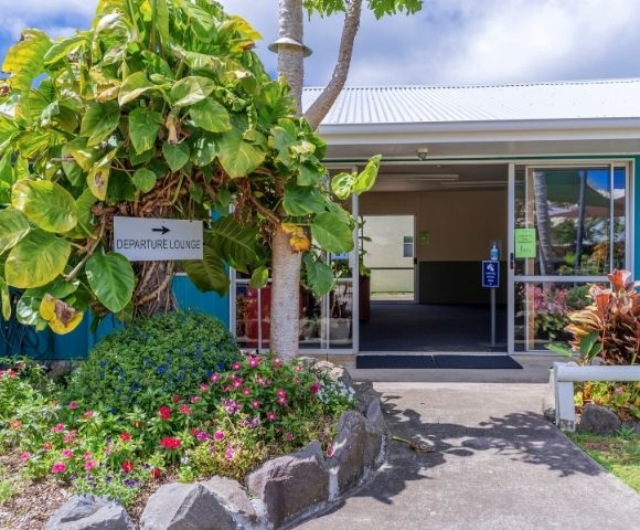A small departure lounge entrance surrounded by lush green plants and colorful flowers on a sunny day, creating a welcoming, tropical atmosphere.