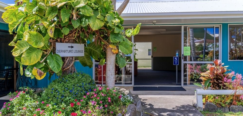 A small departure lounge entrance surrounded by lush green plants and colorful flowers on a sunny day, creating a welcoming, tropical atmosphere.
