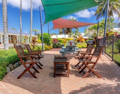 Outdoor patio with wooden chairs around a table, shaded by colorful triangular canopies. Lush greenery and palm trees in the background, under a bright blue sky.