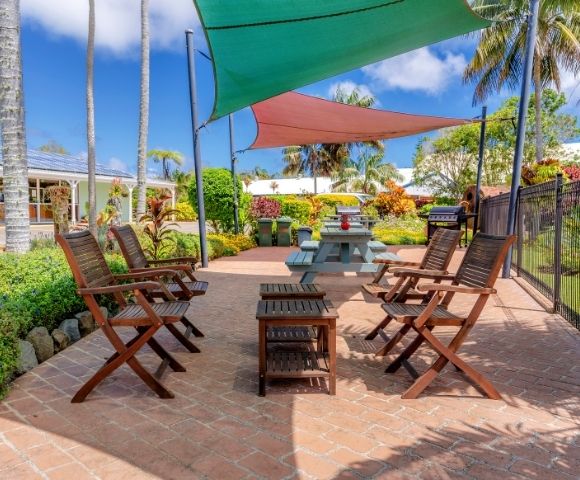Outdoor patio with wooden chairs around a table, shaded by colorful triangular canopies. Lush greenery and palm trees in the background, under a bright blue sky.