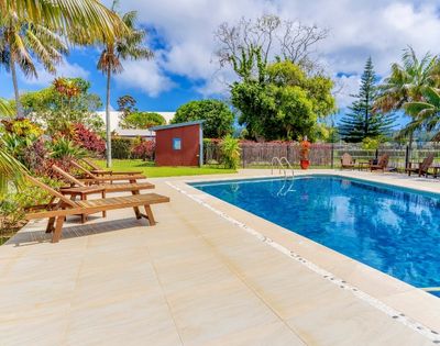 Outdoor pool with clear blue water surrounded by lush trees and palm fronds. Wooden lounge chairs line the deck, creating a serene, tropical vibe.
