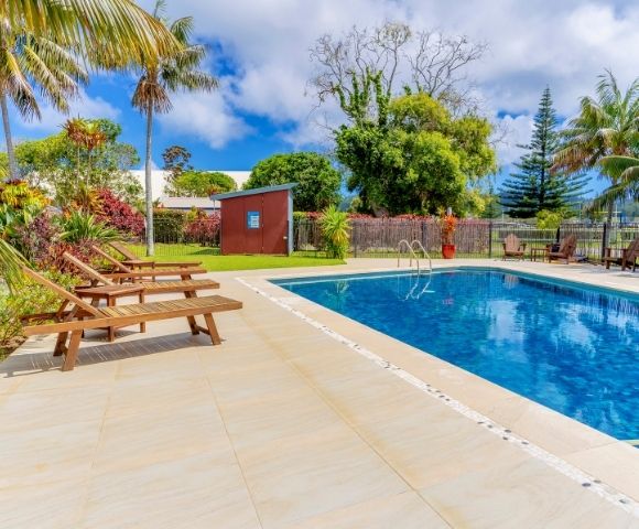 Outdoor pool with clear blue water surrounded by lush trees and palm fronds. Wooden lounge chairs line the deck, creating a serene, tropical vibe.