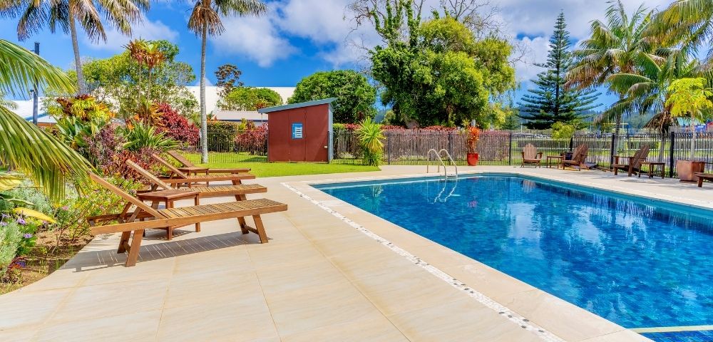 Outdoor pool with clear blue water surrounded by lush trees and palm fronds. Wooden lounge chairs line the deck, creating a serene, tropical vibe.