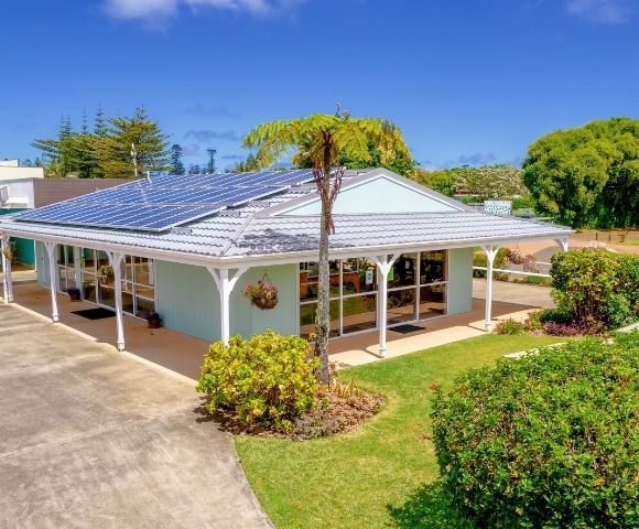 A modern, sunlit house with solar panels on a white roof, surrounded by lush greenery and a clear blue sky, conveying eco-friendly tranquility.