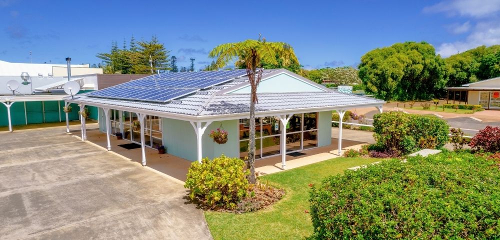 A modern, sunlit house with solar panels on a white roof, surrounded by lush greenery and a clear blue sky, conveying eco-friendly tranquility.