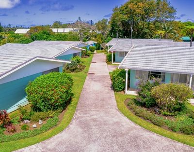 Aerial view of a suburban neighborhood with three small houses, white roofs, light-colored walls, surrounded by lush greenery and a central path. Calm, sunny atmosphere.