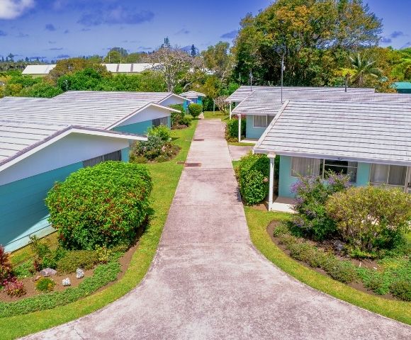 Aerial view of a suburban neighborhood with three small houses, white roofs, light-colored walls, surrounded by lush greenery and a central path. Calm, sunny atmosphere.