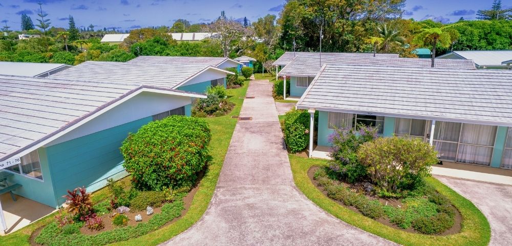 Aerial view of a suburban neighborhood with three small houses, white roofs, light-colored walls, surrounded by lush greenery and a central path. Calm, sunny atmosphere.