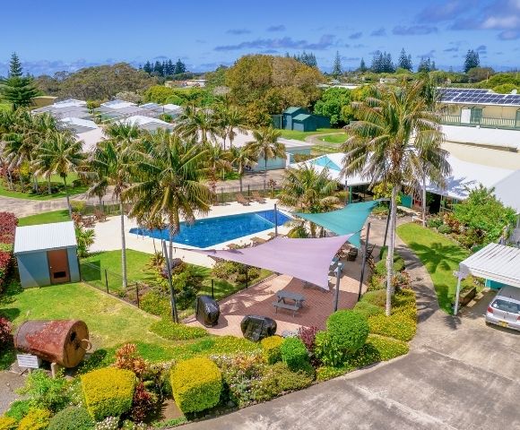 Aerial view of a tropical resort featuring a central swimming pool surrounded by palm trees, colorful shade sails, lush gardens, and various buildings.