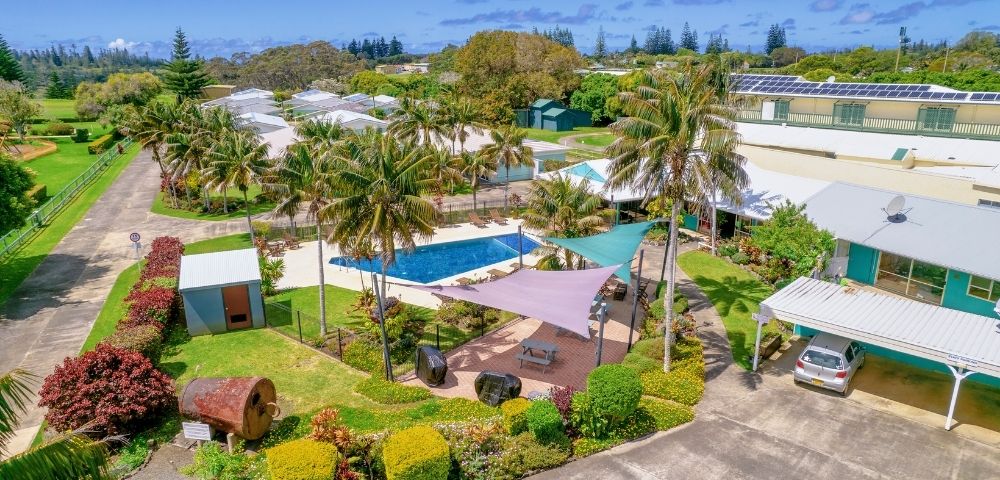 Aerial view of a tropical resort featuring a central swimming pool surrounded by palm trees, colorful shade sails, lush gardens, and various buildings.