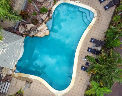 Aerial view of a kidney-shaped pool surrounded by tan tiles, lush greenery, and lounge chairs. The tranquil setting conveys relaxation and leisure.