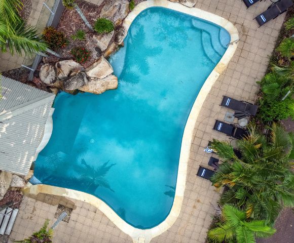 Aerial view of a kidney-shaped pool surrounded by tan tiles, lush greenery, and lounge chairs. The tranquil setting conveys relaxation and leisure.
