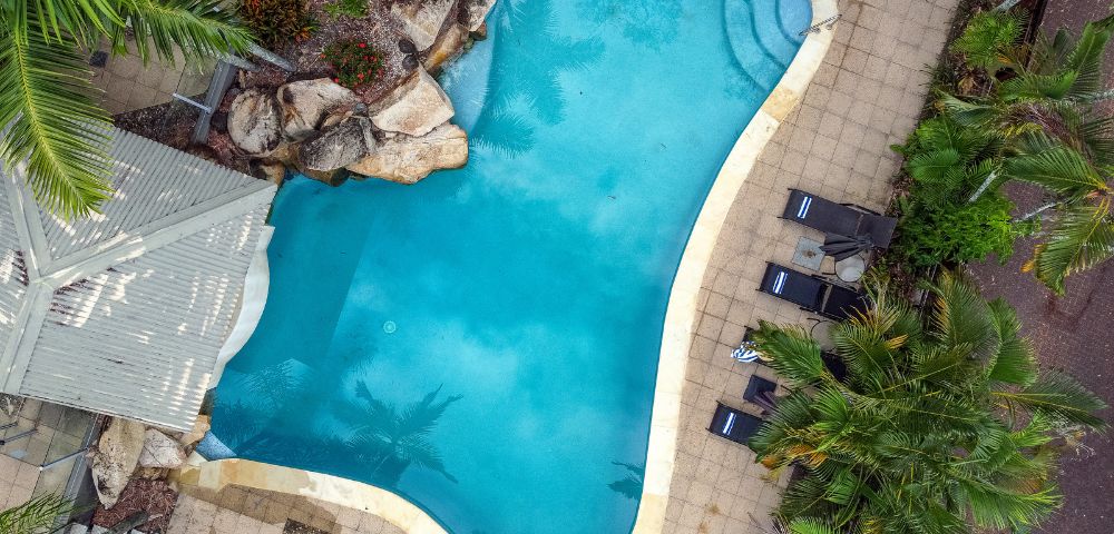 Aerial view of a kidney-shaped pool surrounded by tan tiles, lush greenery, and lounge chairs. The tranquil setting conveys relaxation and leisure.