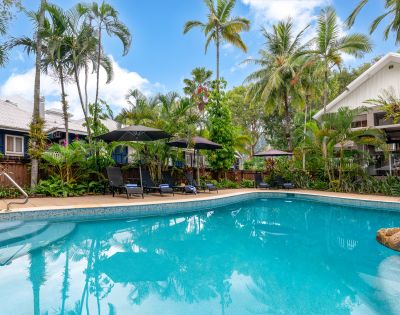 Tropical poolside scene with clear blue water, surrounded by lush palm trees and greenery. Black umbrellas and sun loungers invite relaxation under a bright sky.