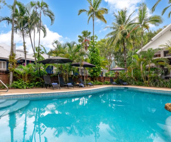 Tropical poolside scene with clear blue water, surrounded by lush palm trees and greenery. Black umbrellas and sun loungers invite relaxation under a bright sky.