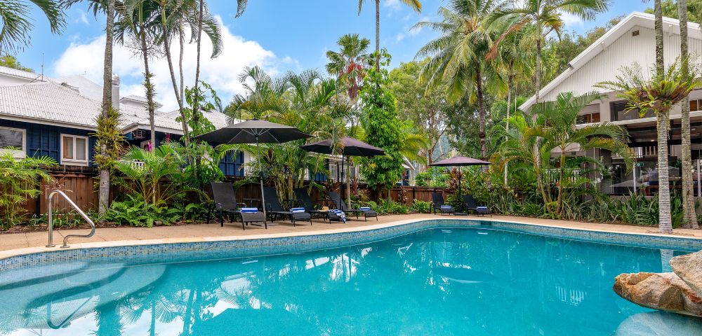 Tropical poolside scene with clear blue water, surrounded by lush palm trees and greenery. Black umbrellas and sun loungers invite relaxation under a bright sky.