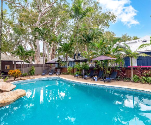 A serene pool scene surrounded by lush trees and tropical plants, with lounge chairs and umbrellas creating a relaxing resort atmosphere under a clear blue sky.