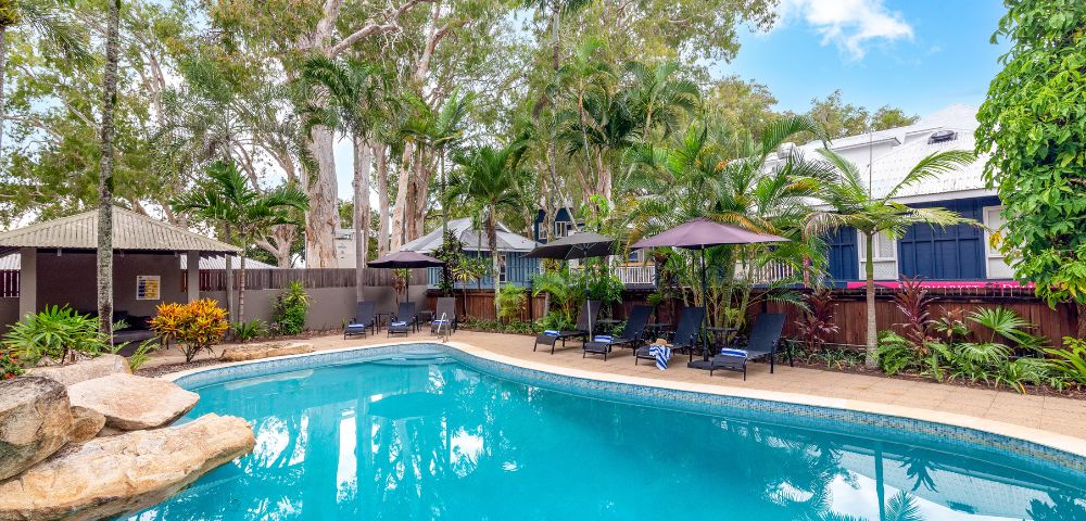 A serene pool scene surrounded by lush trees and tropical plants, with lounge chairs and umbrellas creating a relaxing resort atmosphere under a clear blue sky.