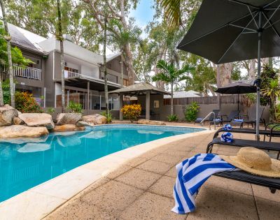 A serene outdoor pool area with lounge chairs, an umbrella, and a striped towel. Surrounded by lush trees and a two-story house, creating a relaxing vibe.