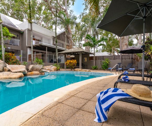 A serene outdoor pool area with lounge chairs, an umbrella, and a striped towel. Surrounded by lush trees and a two-story house, creating a relaxing vibe.