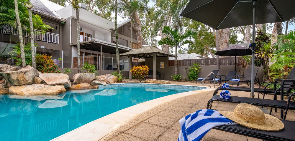 A serene outdoor pool area with lounge chairs, an umbrella, and a striped towel. Surrounded by lush trees and a two-story house, creating a relaxing vibe.