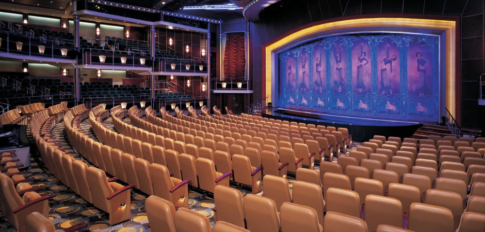 A spacious, elegant theater with curved rows of beige seats facing a vibrant blue stage curtain. The ambiance is warm and inviting, exuding anticipation.