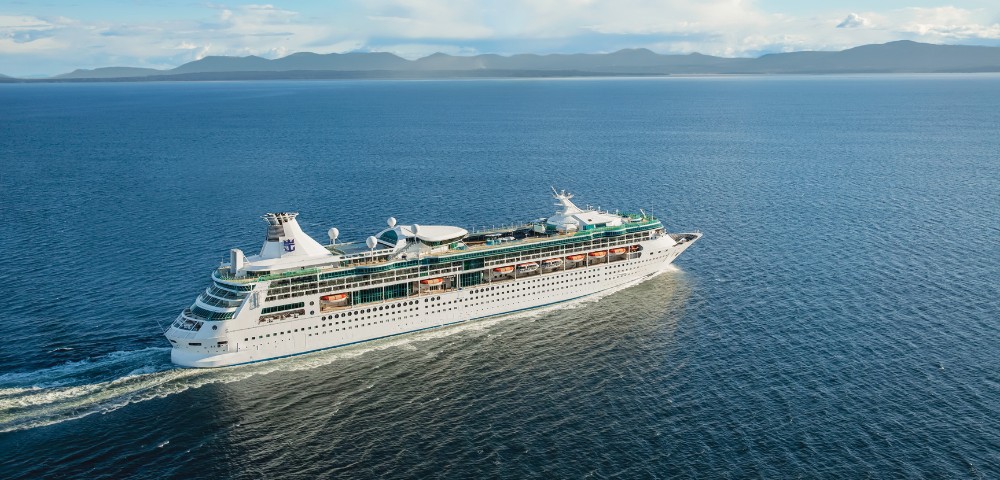 A large white cruise ship sails through calm, blue waters under a bright sky. Lush, green coastline and gentle mountains line the horizon.