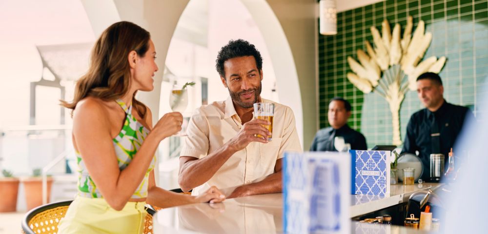 A smiling man and woman enjoy drinks at a stylish bar with a green tiled wall and decorative plant feature. Two bartenders are in the background.