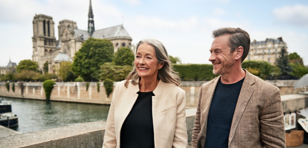 A smiling couple strolls along a riverbank with Notre Dame in the background.