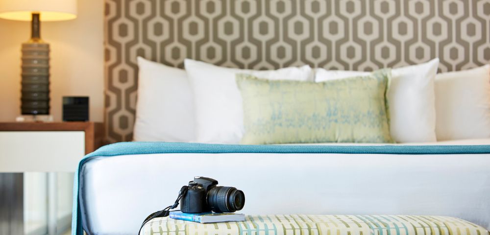A cozy, modern bedroom with geometric wallpaper, a neatly made bed with accent pillows, a bedside lamp, and a camera resting on a patterned bench.
